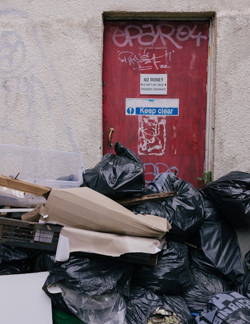 A pile of black bin bags and discarded flattened cardboard boxes is stacked in front of a red metal door embedded in a textured, off-white concrete wall. The bags appear to be full, with some tied at 