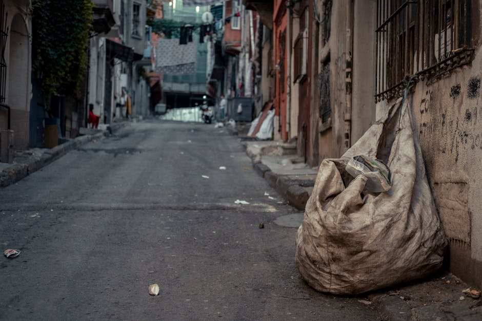 A pile of black bin bags and discarded flattened cardboard boxes is stacked in front of a red metal door embedded in a textured, off-white concrete wall. The bags appear to be full, with some tied at 