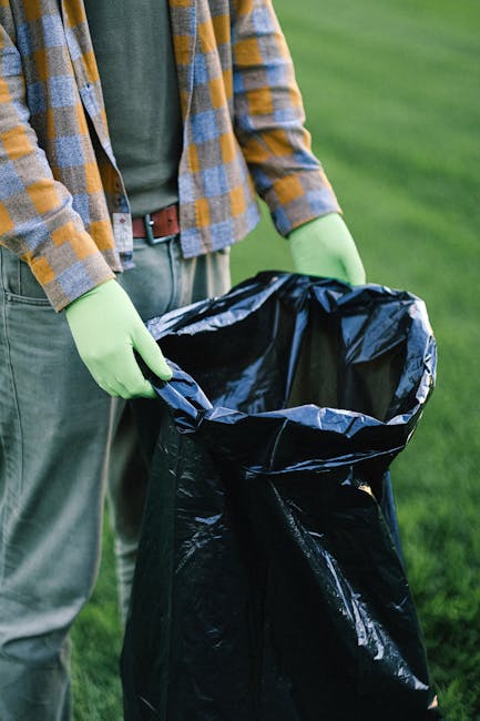 A close-up view of an outdoor rubbish collection site showing a mixture of discarded items including several black garbage bags overflowing with mixed waste, a yellow plastic container lying on the gr